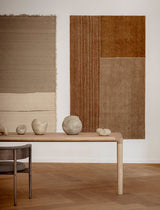 Two abstract rugs on a white wall with a wooden table and stones in the foreground.