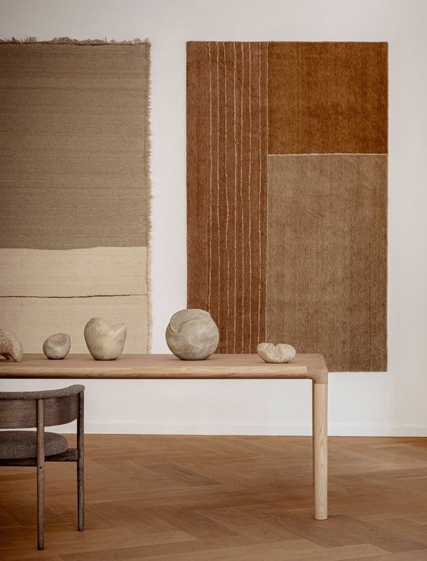 Two abstract rugs on a white wall with a wooden table and stones in the foreground.