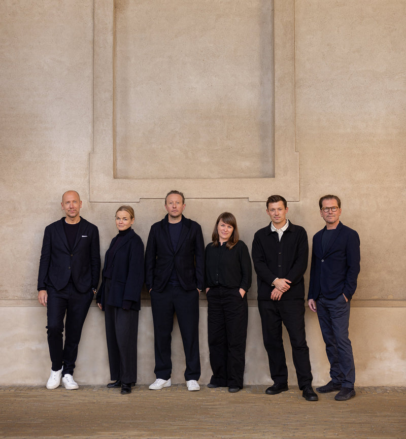 Six people standing in a row against a beige wall.